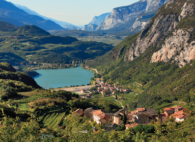 Lago di Santa Massenza. Nell’immagine, il lago da lontano: intorno, l’omonimo borgo e la sua frazione di Vezzano, la montagna con il suo lato roccioso, i prati e i boschi il cui verde si riflette nell’acqua del lago.