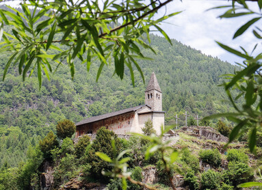Church of Santo Stefano di Carisolo. The church stands atop a granite cliff. Behind, the forest climbs up the side of the mountain: the green of its trees mingles with that of the leaves that, blurred in the foreground, frame the image. On the south-facing side of the church, the one immortalised in the photograph, frescoes can be recognised in the distance. The church has a small stone bell tower and, to its right, three wooden crosses stand out.  